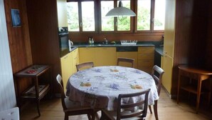 A cozy wooden cabin with a dining area featuring a round table and chairs, a kitchen with a window, and a hanging light fixture. - Chalet am See in St-Prex by Interhome (Saint-Prex)