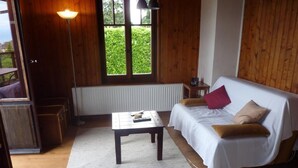 A cozy room with a wooden floor, a white sofa with cushions, a small table, and a window with a view of greenery. - Chalet am See in St-Prex by Interhome (Saint-Prex)