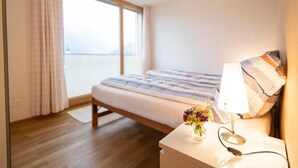Un dormitorio con cama, mesita de noche con lámpara, un jarrón con flores y vistas al mar a través de un ventanal grande.