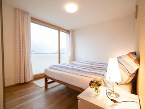 Ein Schlafzimmer mit Bett, Nachttisch mit Lampe, Blumenvase und Meerblick durch ein großes Fenster.