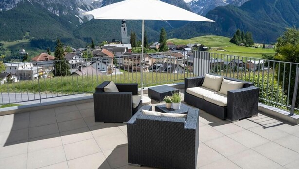 Una terraza con mobiliario de mimbre y una sombrilla blanca, con vistas a un pueblo de montaña con casas y una iglesia.