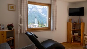 Una habitación con vista a la montaña, una butaca negra reclinable, una estantería de madera, un televisor de pantalla plana y una ventana con cortinas.