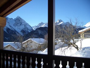 Une vue sur la montagne depuis un balcon en bois, avec des maisons enneigées et des arbres sans feuilles.