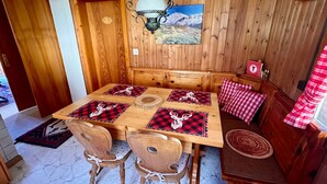Een houten eethoek met een tafel voor vier personen, een bank met geruit kussens, en een schilderij van een berglandschap aan de muur.