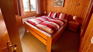 Een slaapkamer met houten lambrisering, een bed, nachtkastje, lamp en raam met gordijnen.