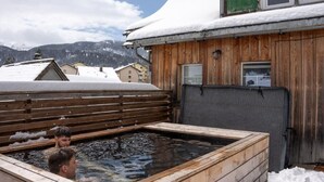 Un bain à remous en bois où se trouvent deux personnes, entouré de bâtiments recouverts de neige et avec des montagnes en arrière-plan.