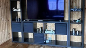 Une pièce aux murs lambrés, avec un grand téléviseur à écran plat posé sur un meuble noir. Des étagères sur ce meuble présentent divers objets, comme des bougies et un panneau lumineux au néon sur lequel on peut lire le mot « Sourire ».