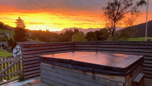 Un bain à remous avec une terrasse en bois et une vue sur un coucher de soleil se déclinant sur un paysage vallonné.