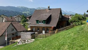 Un chalet traditionnel avec un toit en pente, une clôture en bois et un mur de pierres, situé dans un cadre rural avec des montagnes en arrière-plan.
