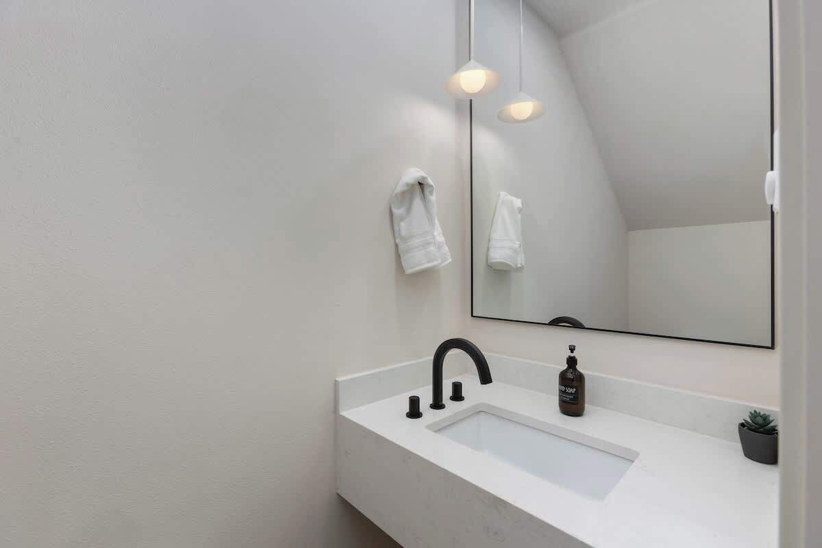 Une salle de bain moderne avec un lavabo blanc, un robinet noir et un grand miroir. Deux serviettes sont accrochées au mur et une petite plante en pot est posée sur le comptoir.
