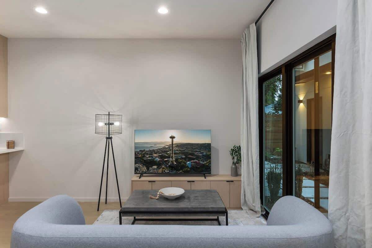 Ein modernes Wohnzimmer mit einem großen Flachbildfernseher an der Wand, einer Stehlampe, einem Esstisch mit Schüssel und Blick auf eine Stadtlandschaft durch ein großes Fenster.