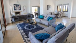 A spacious living room with a grey sofa set, a coffee table, and a chandelier. There is a dining area with a table and chairs, and a fireplace with a mantel. - Villa mit Blick auf die Berge und Whirlpool by Interhome (Kathikas Village)