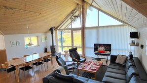 Un vaste espace de vie doté d’un plafond en bois, d’un grand canapé, d’une table à manger avec des chaises, d’un téléviseur et d’une vue sur l’extérieur.