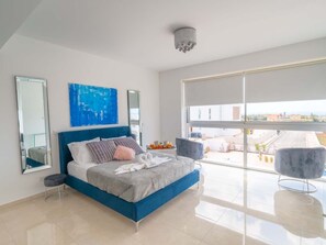 Ein modernes Schlafzimmer mit einem großen Bett, zwei Stühlen, einem blauen Kopfteil, einem großen Fenster und Blick auf die Stadt.