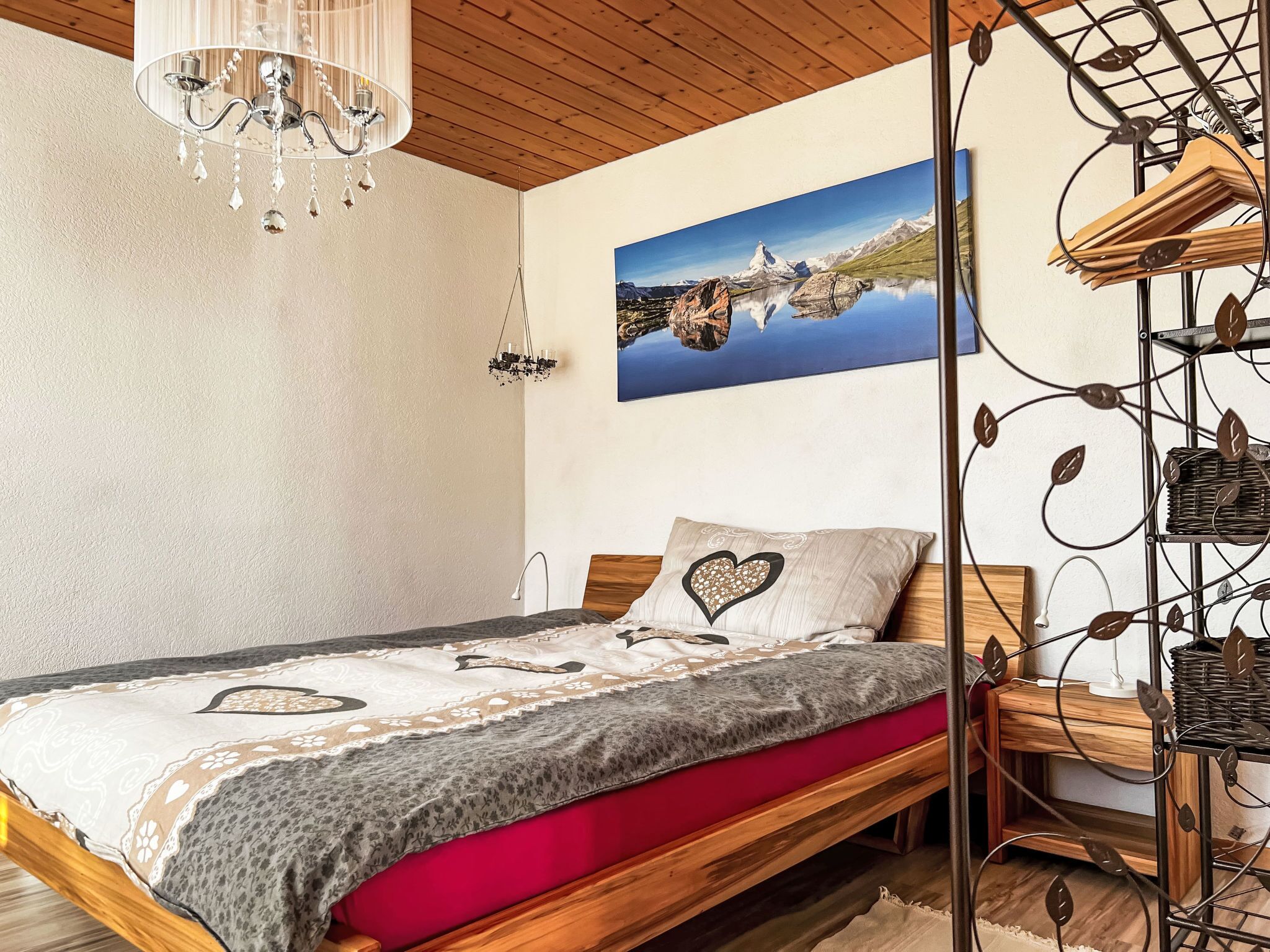 Une chambre à coucher avec un lit en bois, un lustre, un tableau représentant un paysage de montagne et un porte-vêtements fixé au mur.