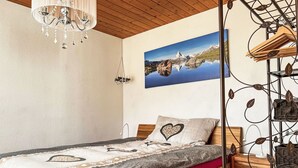 A bedroom with a wooden bed, a chandelier, a painting of a mountain landscape, and a wall-mounted rack with clothes.