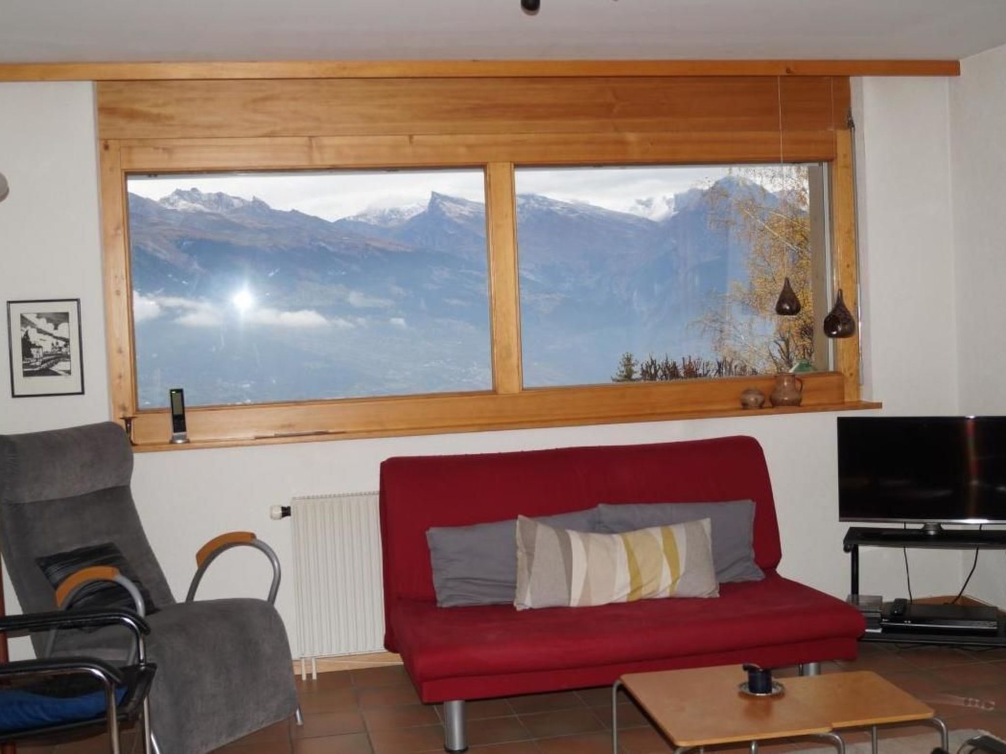 A living room with a red sofa, a TV, a window with a mountain view, a radiator, and a chair.