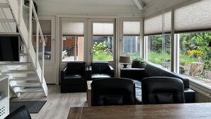 A modern living room with a wooden dining table, black leather sofas, a staircase, and large windows offering a view of a garden.
