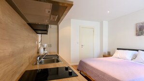 Kompakti hotellihuone, jossa on pieni keittiönurkkaus, violetilla päiväpeitteellä varustettu sänky ja valkoinen ovi.