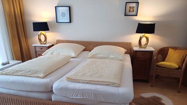 Een tweepersoonsbed met witte lakens, twee nachtkastjes met lampen, een rieten stoel met een geel kussen, en een ingelijst kunstwerk aan de muur.
