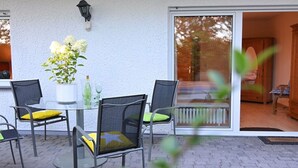 Een terras buiten met een glazen tafel, vier stoelen, een potplant en een schuifpui die naar een binnenruimte leidt.