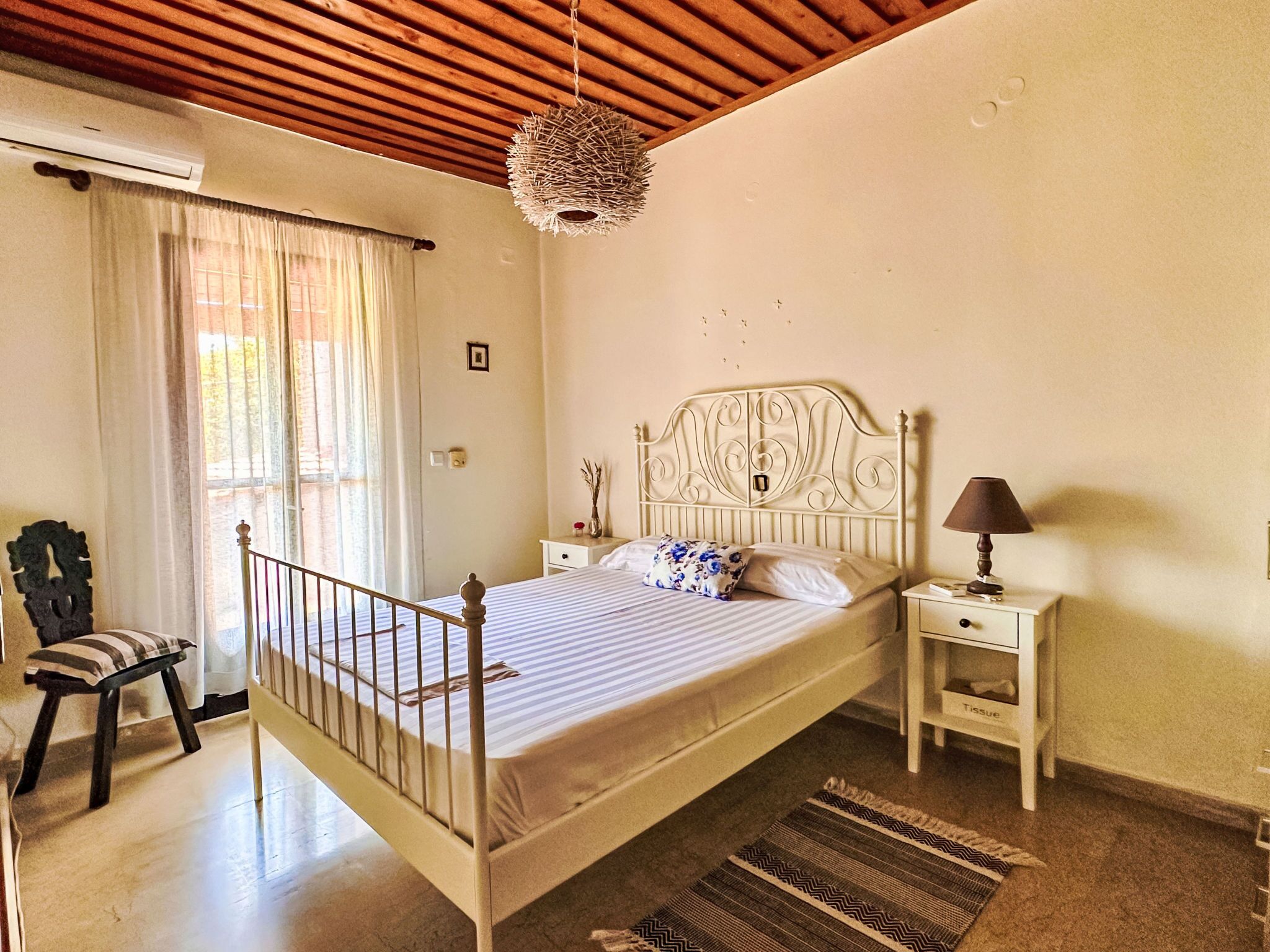 Un dormitorio con cama, mesitas de noche, una silla, una lámpara colgante y una ventana con cortinas.
