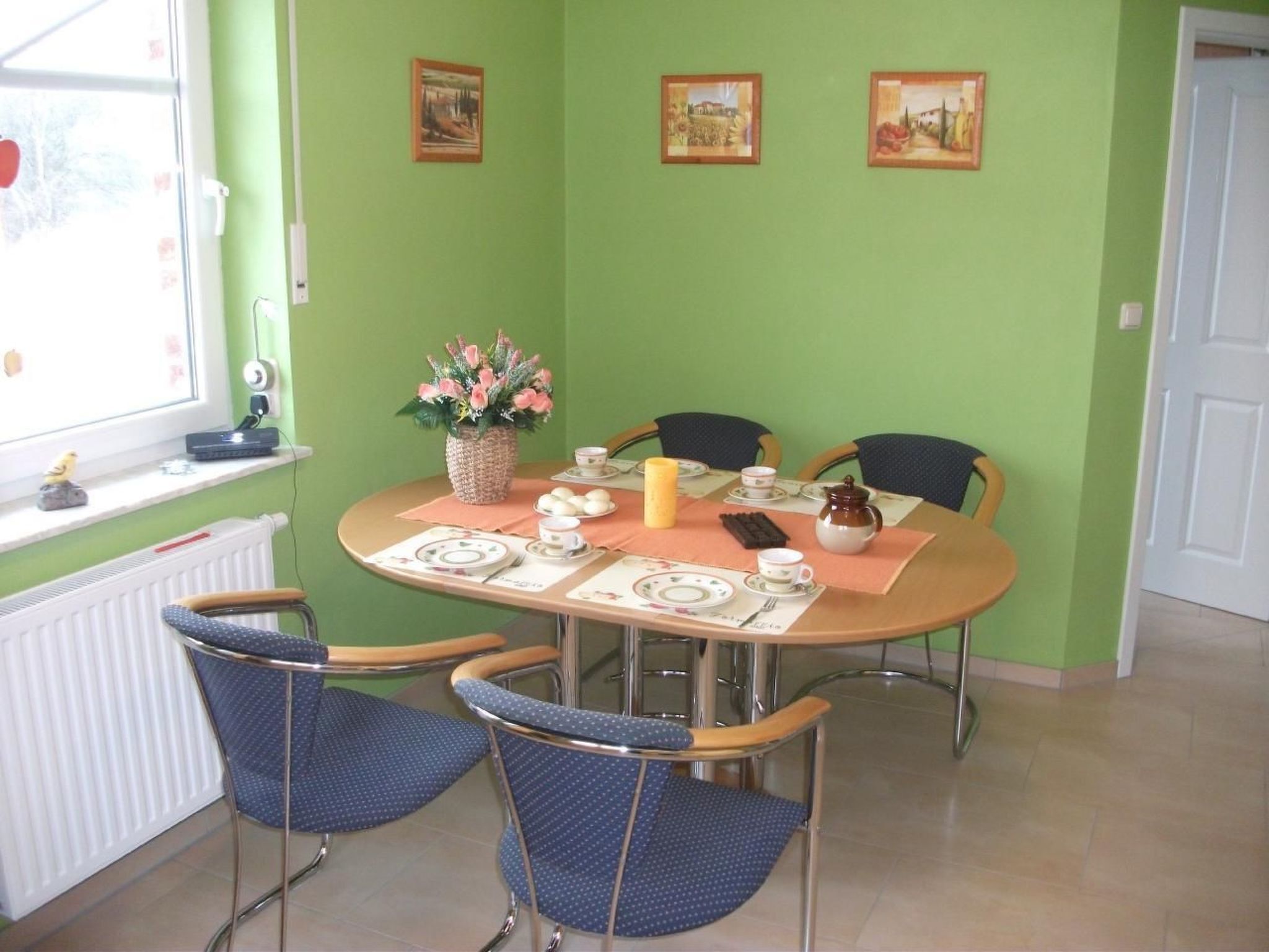 Un coin repas avec une table ronde dressée pour quatre personnes, entourée de chaises bleues. Trois tableaux encadrés sont accrochés au mur au-dessus de la table.