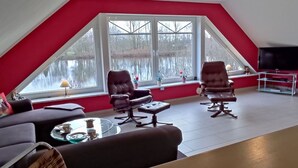 Una sala de estar moderna con techo inclinado, paredes rojas y grandes ventanas con vistas a un cuerpo de agua. Hay dos sillones de cuero, una mesa de centro de vidrio y un sofá.