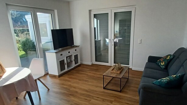 Een moderne woonkamer met een flatscreen televisie op een wit kastje, een grijze bank met decoratieve kussens, een houten salontafel, en een eethoek met een witte tafel en stoelen.