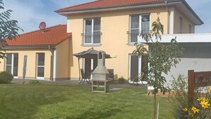 Een huis met twee verdiepingen, voorzien van een terras met een barbecue en een kleine tuin met gele bloemen.
