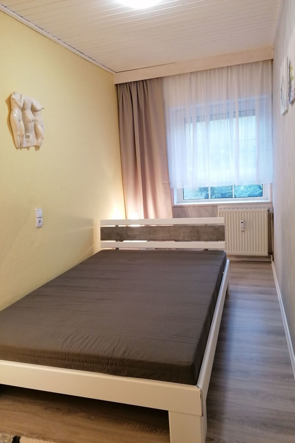 Ein Schlafzimmer mit einem Einzelbett, einem Heizkörper, einem Fenster mit Vorhängen und einer Wandskulptur.