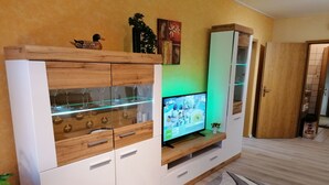 Ein modernes Wohnzimmer mit einem Fernseher auf einem Holzständer, einer Glasvitrine mit Weingläsern und einem gemusterten Teppich.