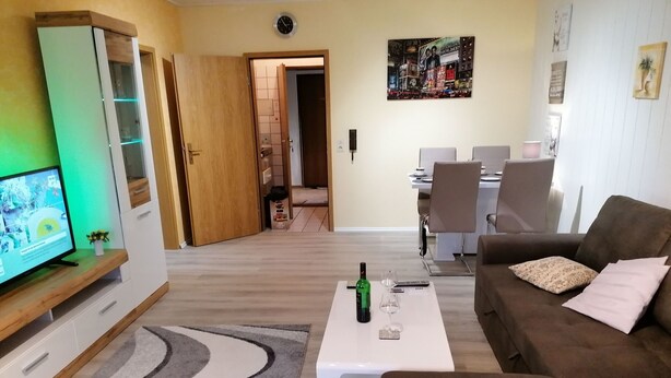 Ein Wohnzimmer mit Fernseher, einem Couchtisch mit Flasche und Gläsern, einem braunen Sofa und einem Essbereich im Hintergrund.