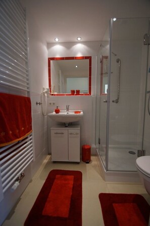 Une salle de bain avec une douche à parois de verre, un lavabo blanc avec un miroir orné de carreaux rouges et des tapis rouges sur le sol.