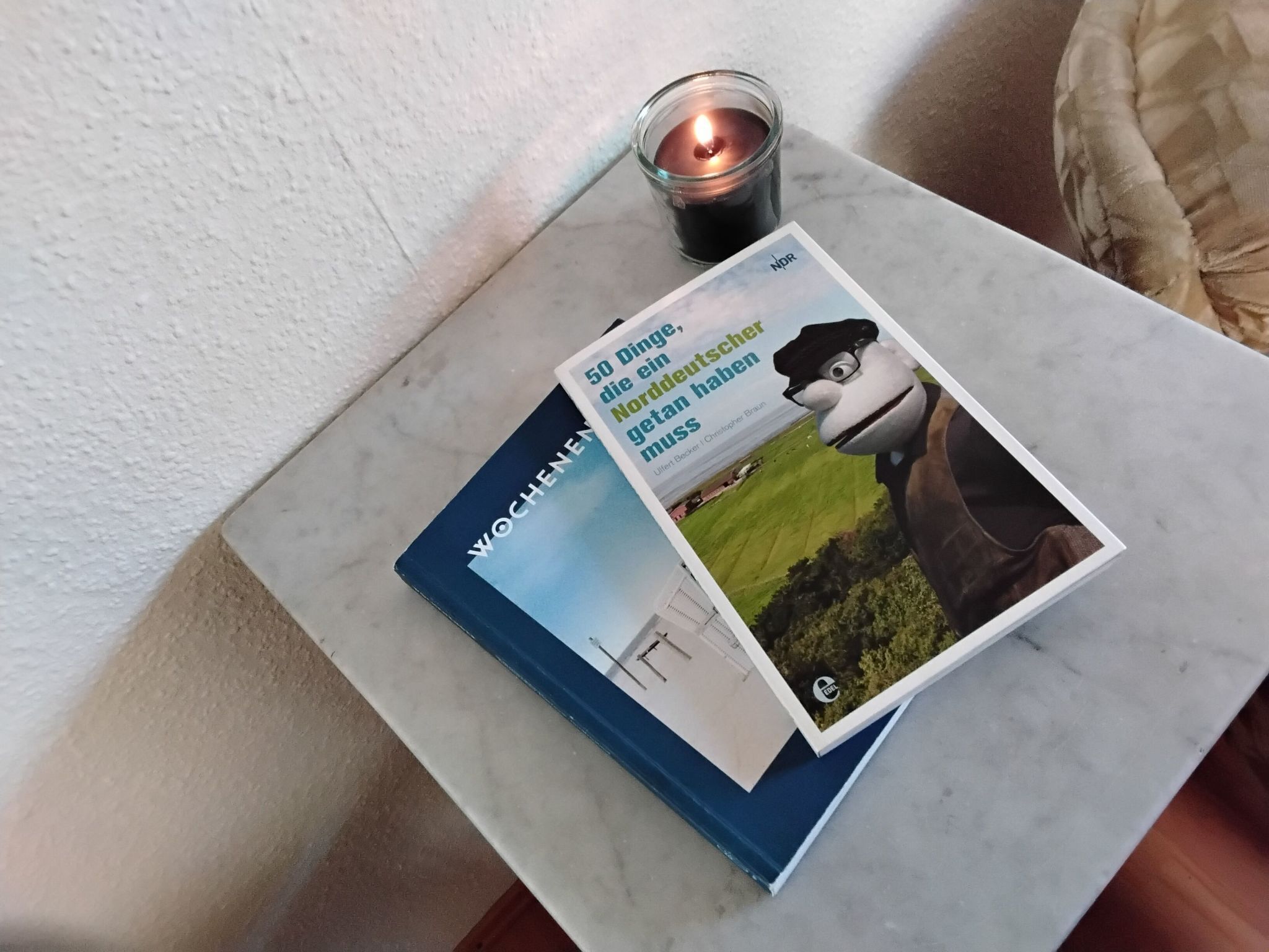 Ein Buch mit dem Titel „50 Dinge, die Deutschland haben“ auf einem Marmortisch neben einer brennenden Kerze.