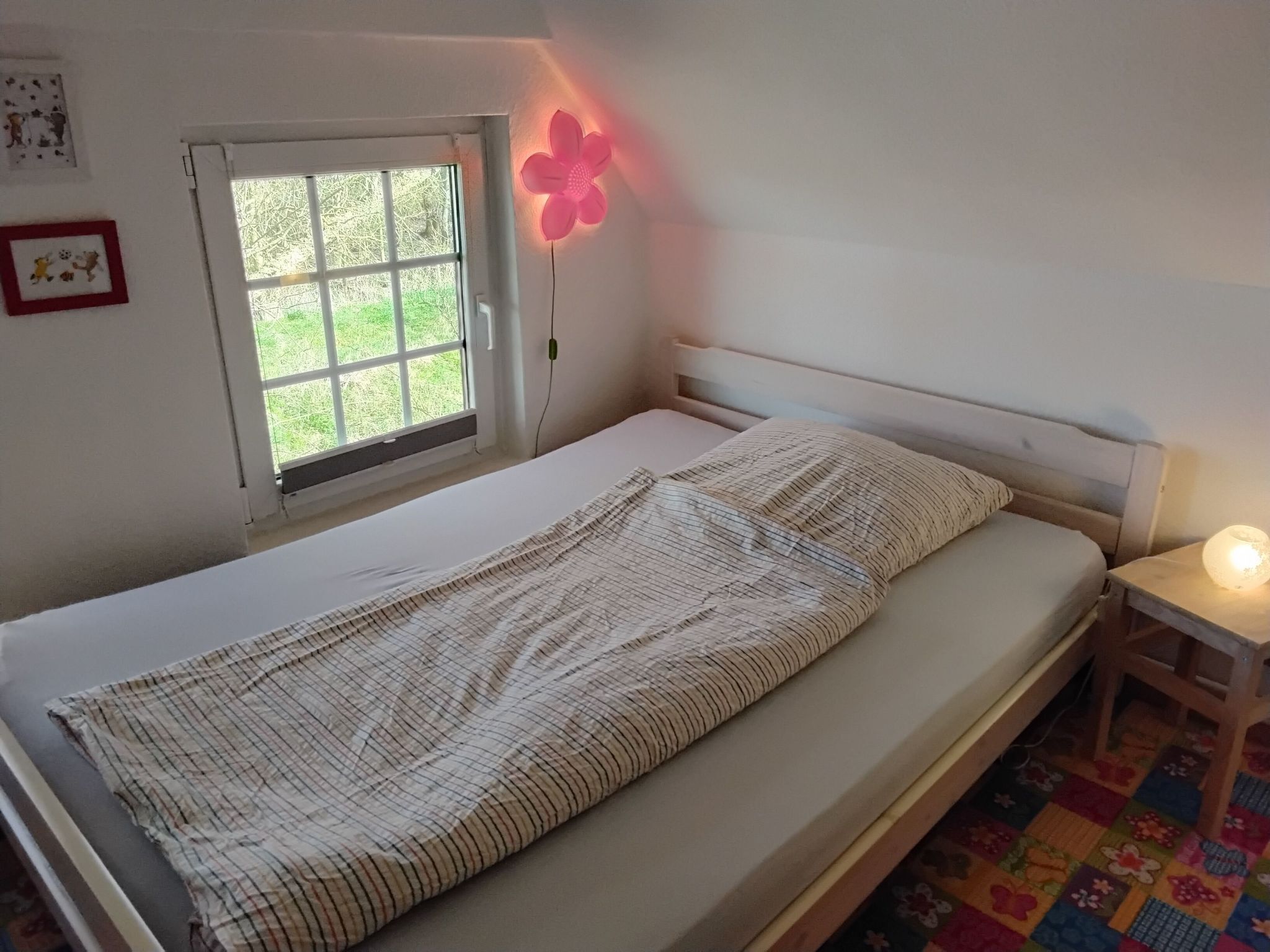 Un lit simple avec une couverture à rayures, une fenêtre donnant sur un paysage verdoyant, une lampe en forme de fleur rose et une petite table de chevet avec une lampe.