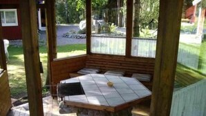 A covered outdoor seating area with a stone table and benches, surrounded by a wooden fence and trees.
