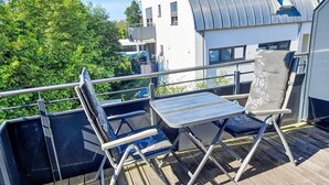 Eine Terrasse mit zwei Stühlen und einem kleinen Tisch, mit Blick auf einen Garten und ein modernes Gebäude mit einer gewölbten Dachform.