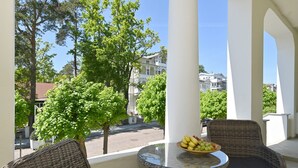 Un balcone con sedie in vimini e un tavolo, una ciotola di frutta e vista su edifici e alberi.