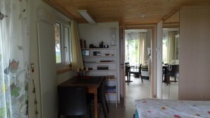 Habitación pequeña con cama, escritorio y silla. Tiene una ventana con cortinas, una estantería con varios objetos y una puerta que da a otra habitación.