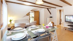 Un coin repas avec une table en verre dressée pour quatre personnes, des poutres en bois au plafond et un téléviseur en arrière-plan.