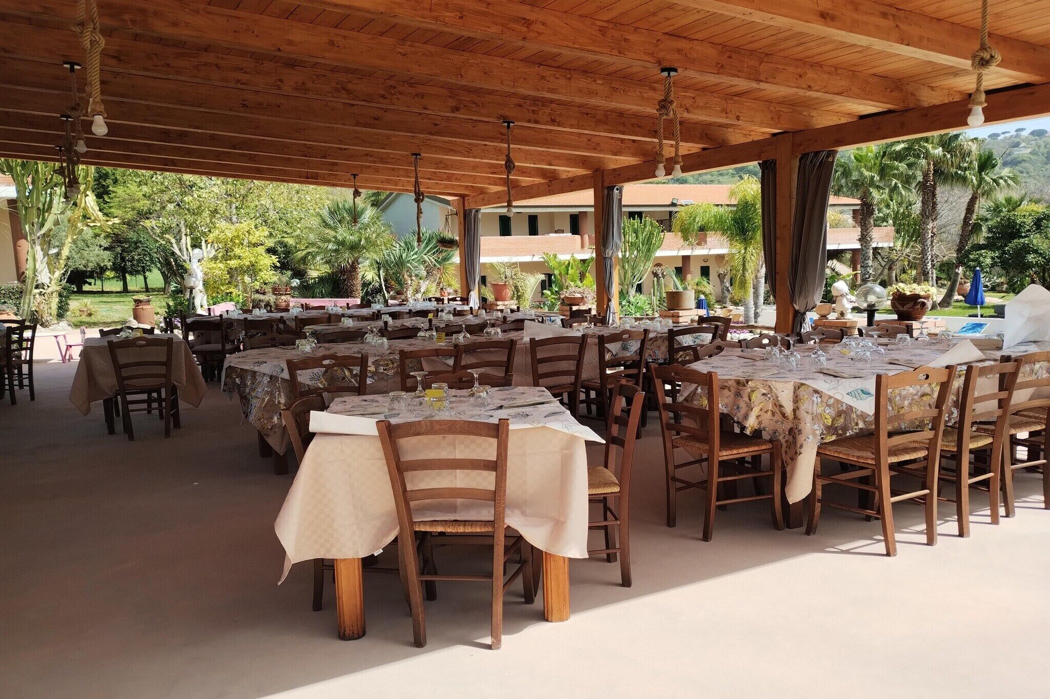 Area pranzo all'aperto con diversi tavoli e sedie, coperta da un tetto in legno, immersa nel verde e con una piscina.