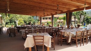 Un área de comedor al aire libre con varias mesas y sillas, cubierta por un techo de madera, rodeada de abundante follaje y una piscina.