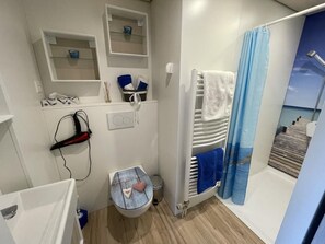 Casa de banho compacta com chuveiro, sanita e uma pequena pia. Existem prateleiras acima da sanita e um porta-toalhas com uma toalha branca.