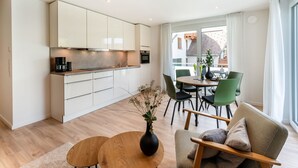 Uma cozinha moderna com armários brancos, um espaço de refeições com cadeiras verdes e uma grande janela com vista para edifícios.