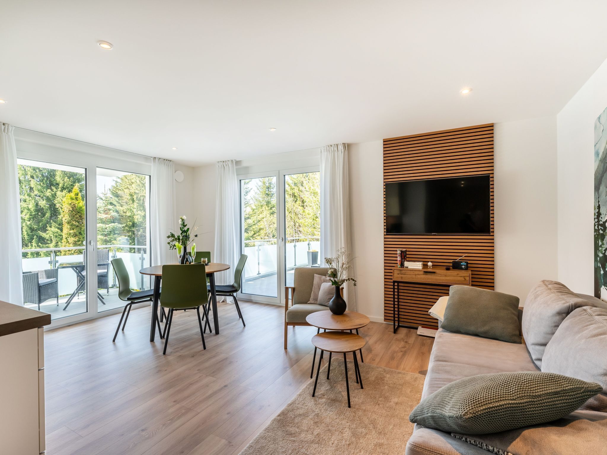 A modern living room with a sofa, a TV mounted on a wooden panel, a dining area with chairs and a table, and large windows offering a view of trees.
