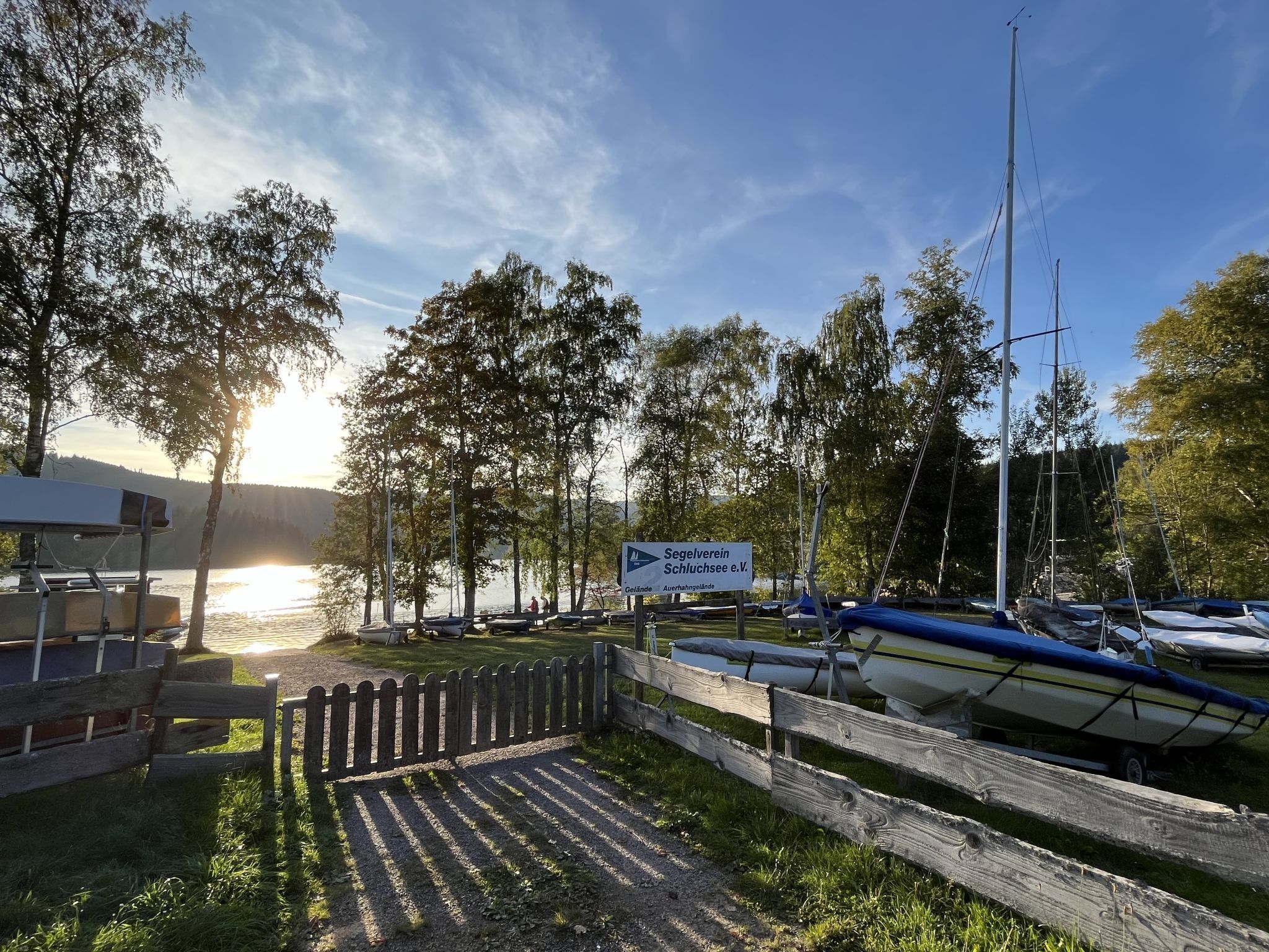 Ein Yachthafen am See mit vertäuten Booten, einer Holzlattenzäunung und einem Schild mit der Aufschrift 'Segelverein'.