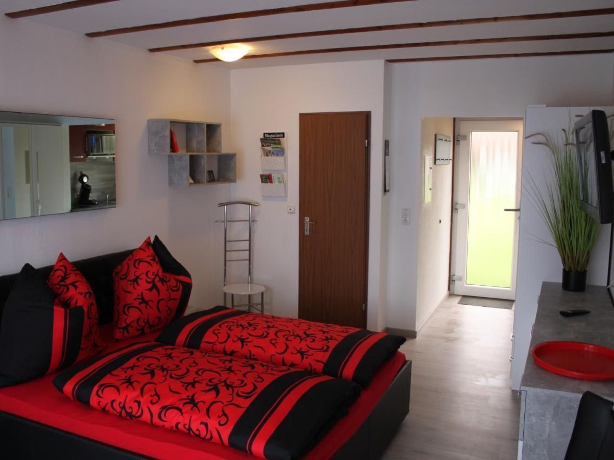 Een moderne slaapkamer met een bed, een spiegel, een klein plankje en een deur naar een gang.