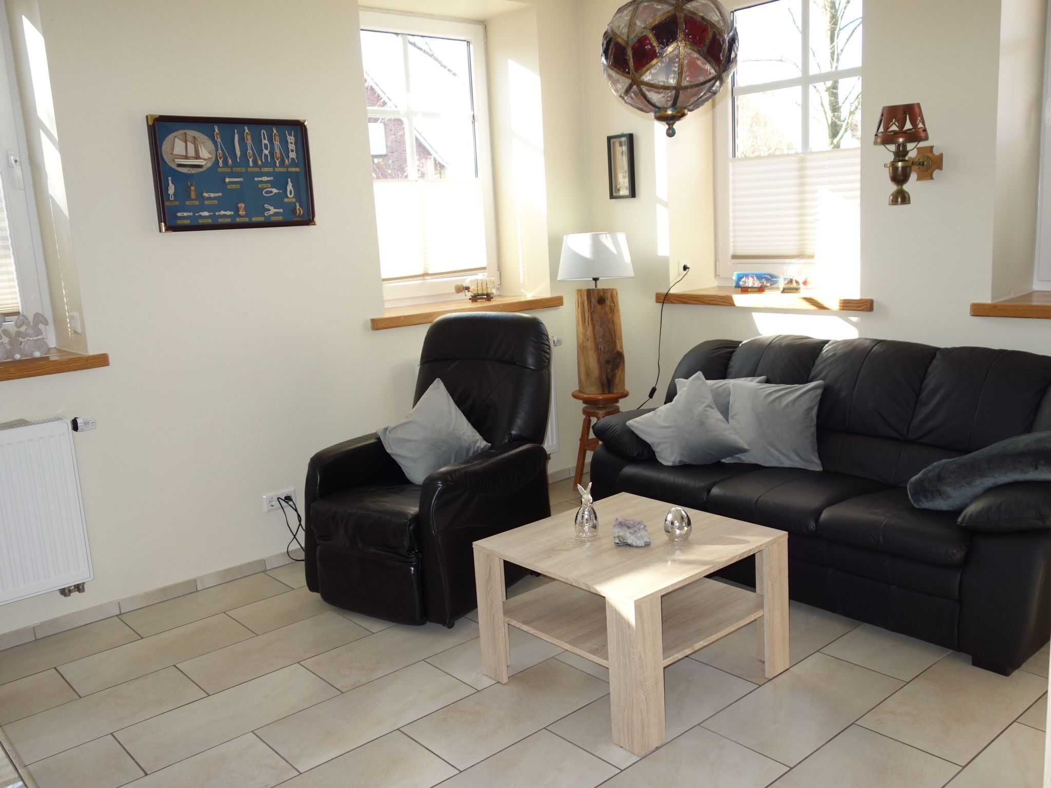 A living room with a black leather sofa, a wooden coffee table, and a black leather armchair. There is a wall-mounted lamp and a framed picture on the wall.