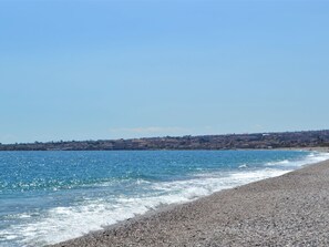 A coastal beach with pebbly shore, clear blue sea, and a distant town. - Saras Terrasse by Interhome (Mascali)